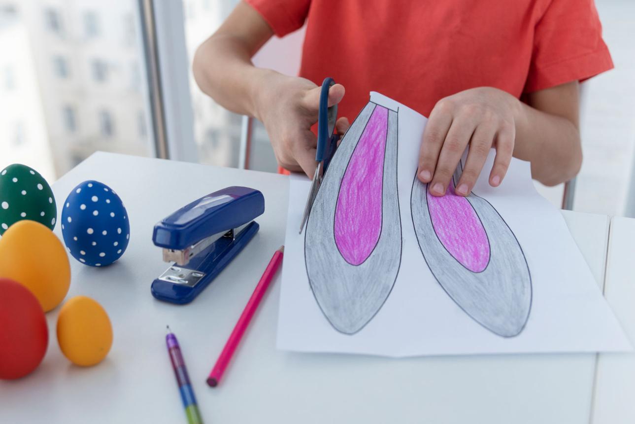 Boy hands cut out easter bunny ears with scissors, colored easter eggs are near on table.