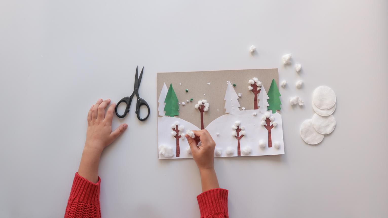 Child's hands creating a winter landscape craft with cotton balls and paper