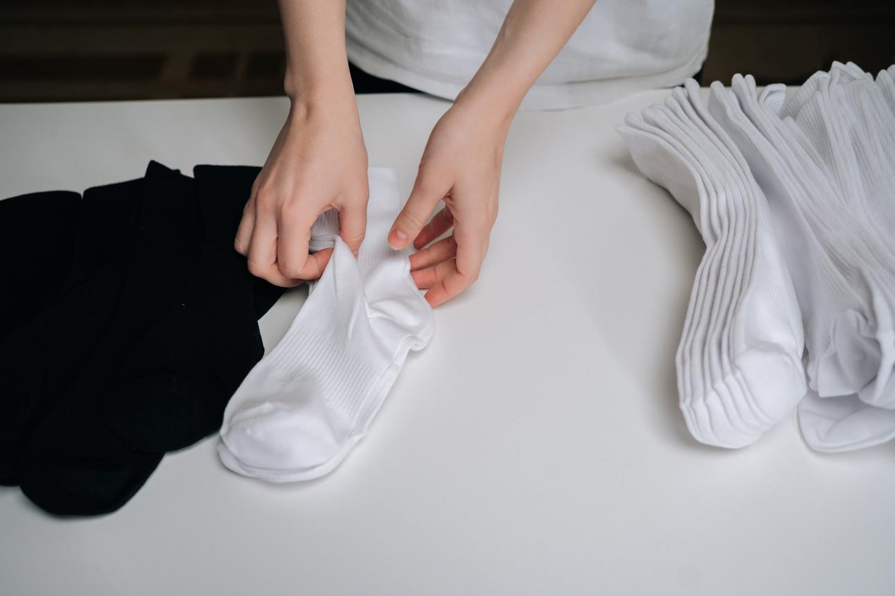 Close-up of hands folding pair of freshly laundered white socks on clean white surface, preparing for organized storage alongside stack of other folded white socks and pile of black socks